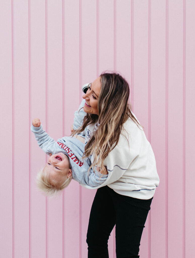 Mum dips toddler son upside down while laughing in front of pink wall