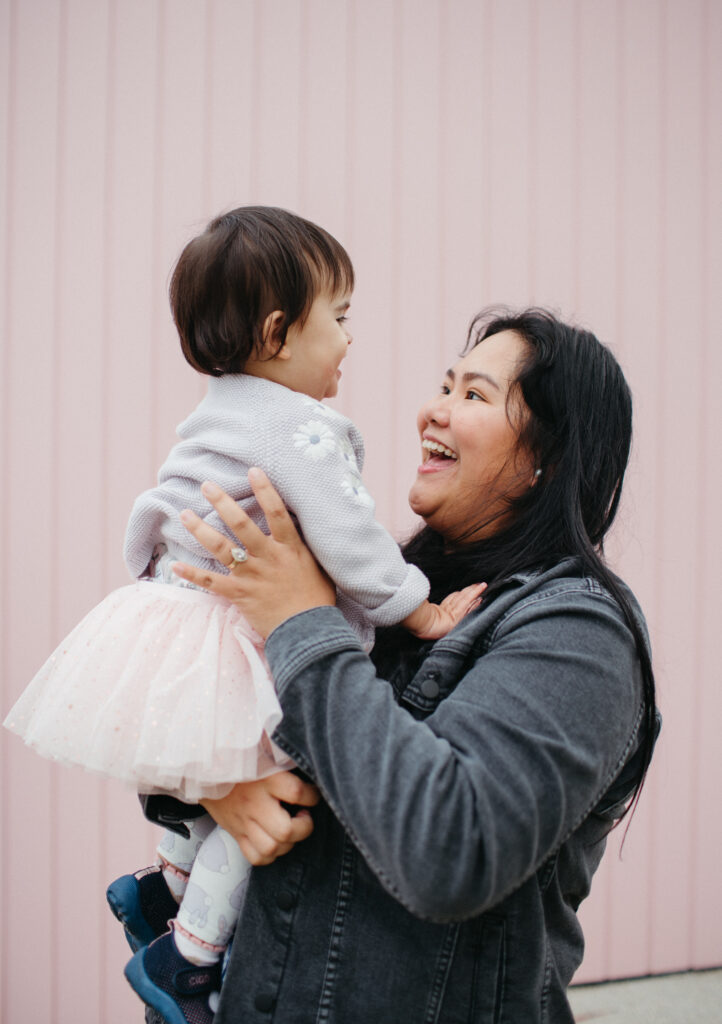 mum lifts her baby girl in the air. Little girl is wearing a tutu in front of a pink wall