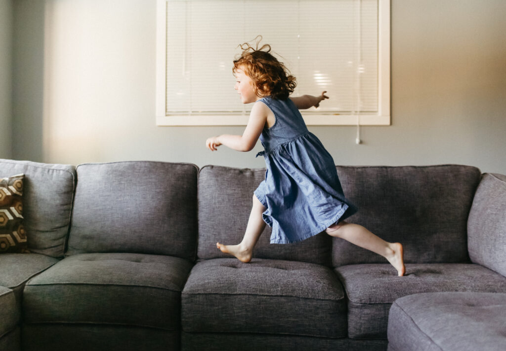 Little girl wearing blue dress runs across couches.