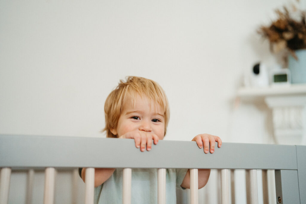 Cute toddler looks out from over his crib bars.