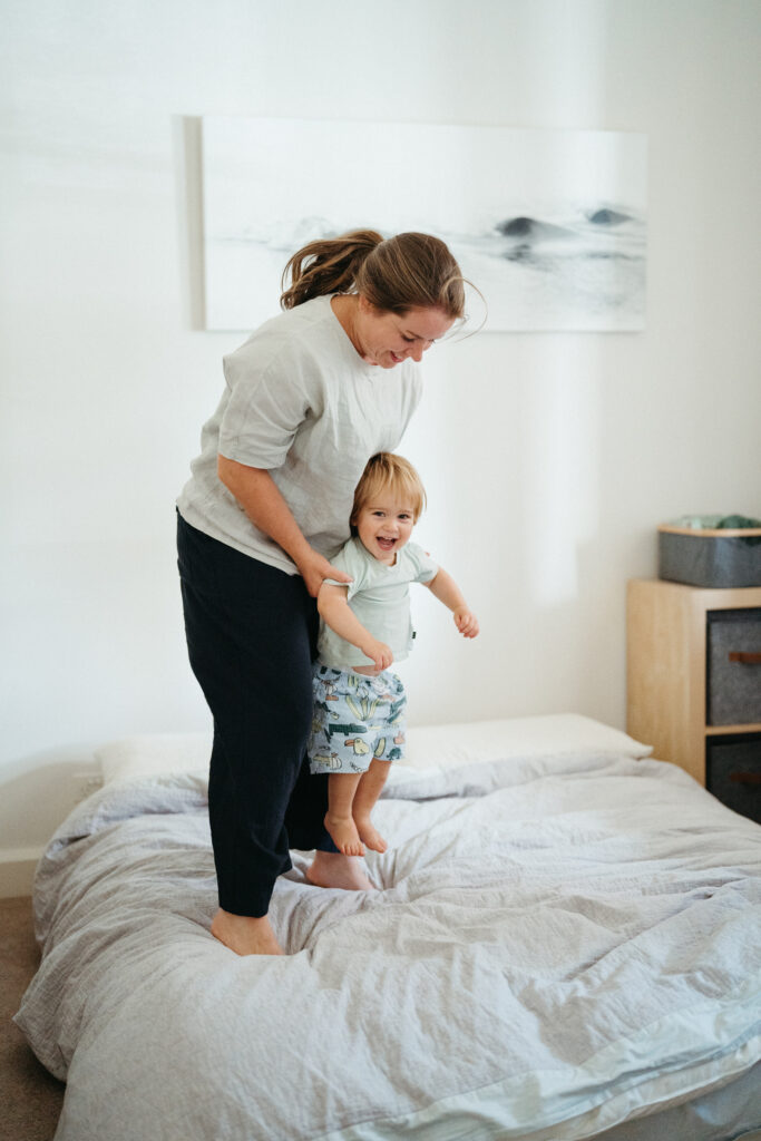 Mum jumps on the bed with her toddler son.